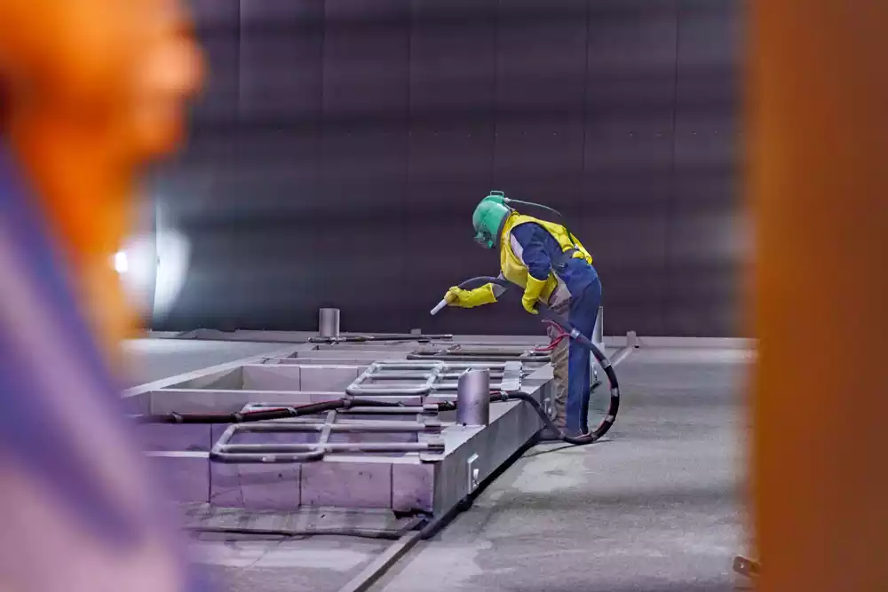Worker in protective gear engaged in abrasive blasting of a large steel frame.