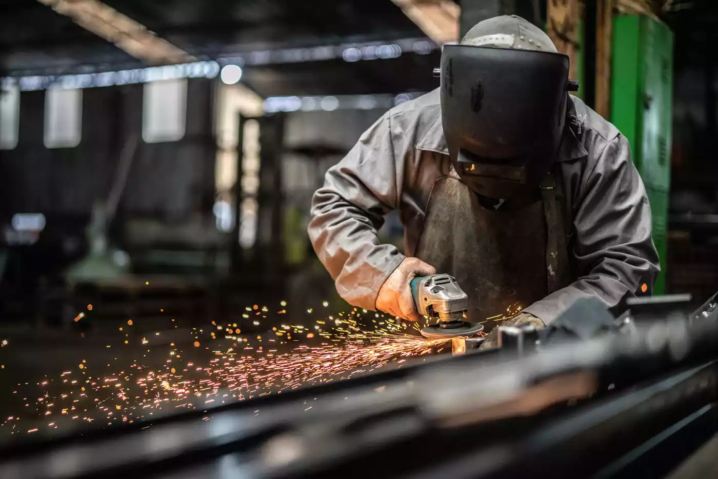 A man in protective gear grinding a large steel part in a workshop, generating visible sparks.