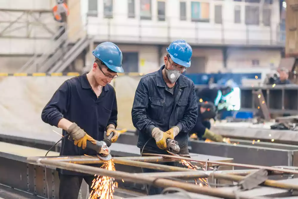 Two men on a construction site wearing dark blue work coveralls and light blue hard hats grinding a large metal frame using manual tools, generating visible sparks.