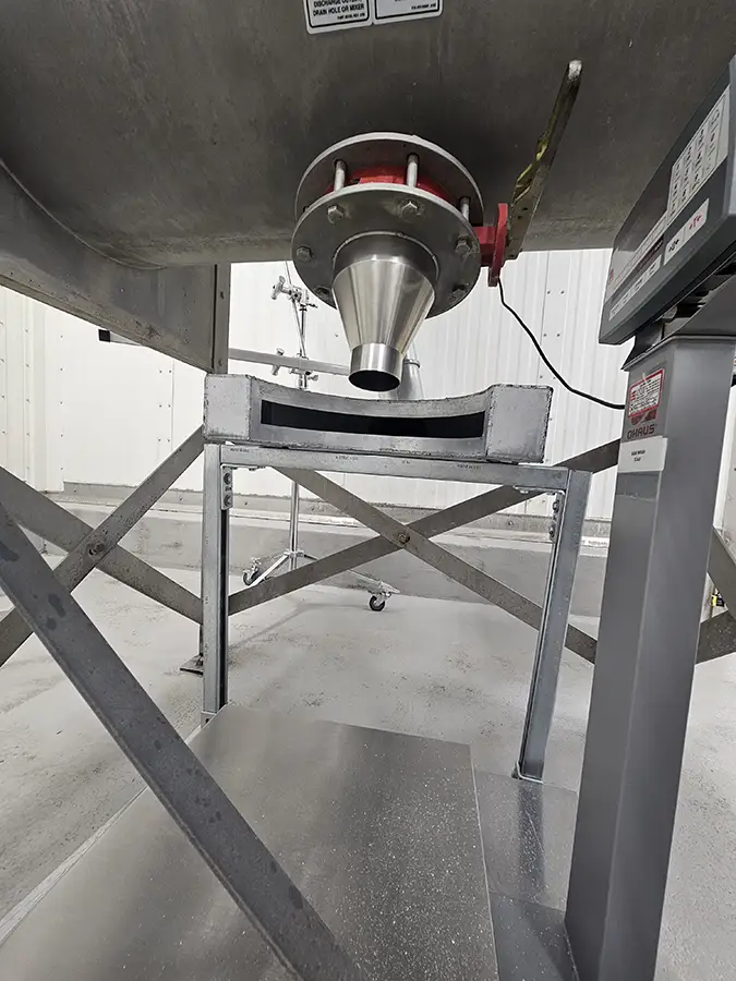Stainless steel powder discharge outlet positioned above an industrial weighing scale in a processing facility.