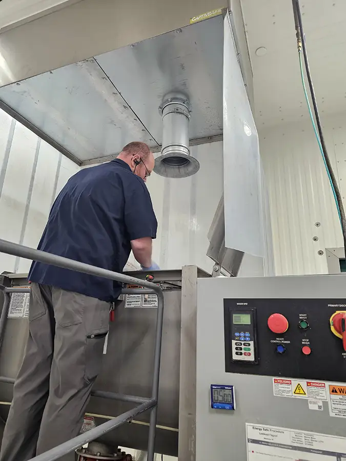 Worker operating an industrial mixer beneath a ventilation hood and dust collection system in a manufacturing facility.